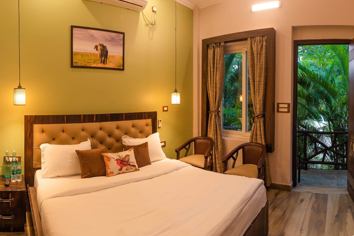 A hotel room with a large bed, a nightstand with bottles, a chair, and a window with curtains.