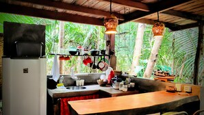 Une cuisine rustique avec une table en bois, des ustensiles de cuisine suspendus et une vue sur une végétation luxuriante.