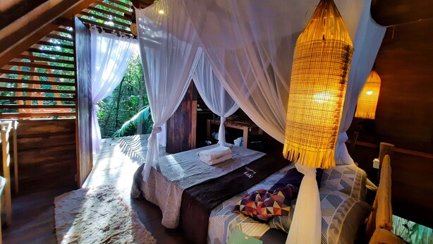 Une chambre à coucher chaleureuse et rustique, dotée d’un lit à baldaquin, d’un luminaire suspendu en fibres tressées et offrant une vue sur une végétation luxuriante.