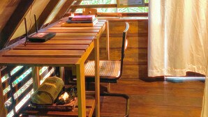 Une chambre mansardée avec un plafond en pente, des poutres en bois, une fenêtre donnant sur la verdure, une petite table avec une chaise et un lit avec une tête de lit.