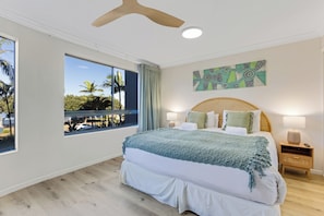 Relax in a spacious room with a large bed, ceiling fan, and wall-mounted artwork, offering a view of palm trees and a pool.