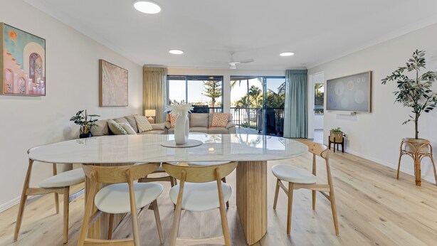 A modern living room with a round glass-top dining table, wooden chairs, a sofa, and a large window offering a view of the outdoors. - Riverdance Apt 6  (Mooloolaba)