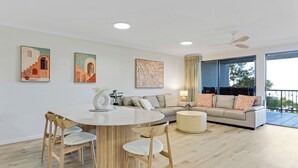 A modern living room with a round dining table, wooden chairs, a sofa, and a large window with a view. - Riverdance Apt 6  (Mooloolaba)