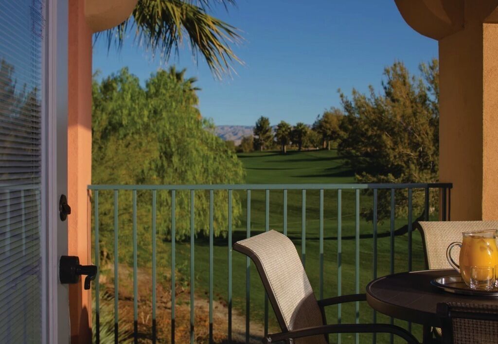 A balcony with a view of a golf course, a chair, and a table with a drink.