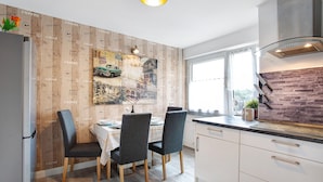 A modern kitchen with a dining area, featuring a coffee-themed wallpaper, a vintage car painting, and a brick accent wall. - Coastal Love Apartment – Close to Park & City (Wilhelmshaven)