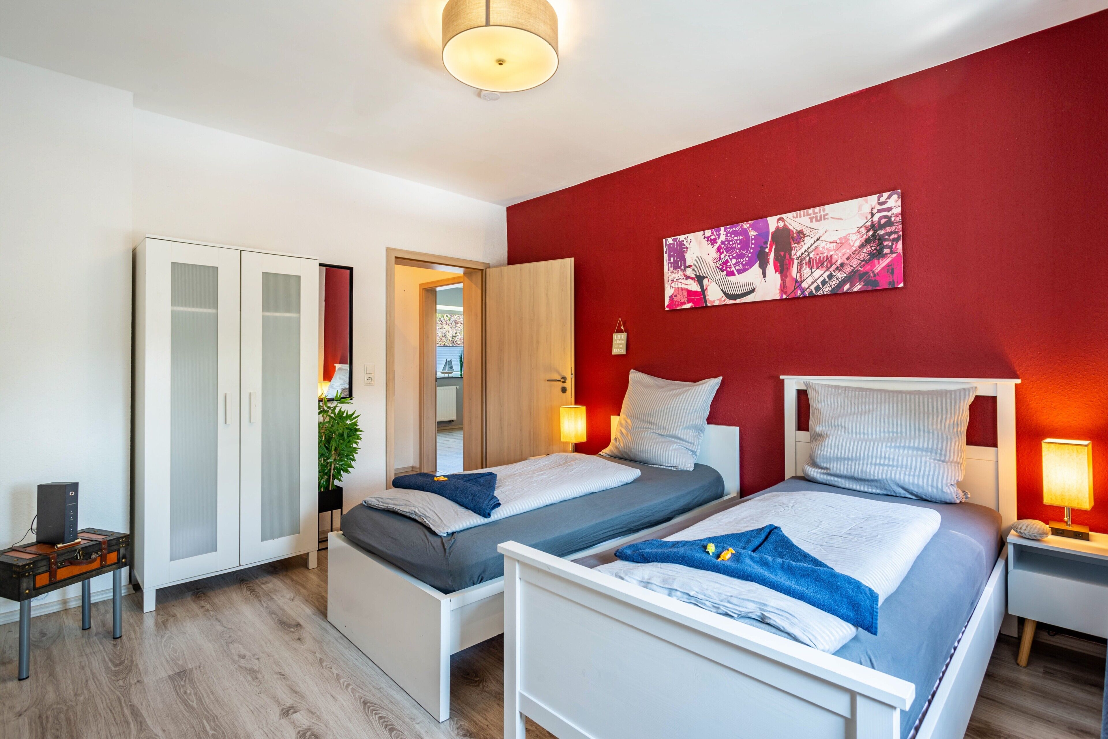 A bedroom with two beds, a wardrobe, a desk, and a red accent wall.
