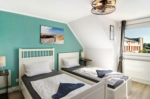 A room with two beds, a view of buildings, a picture on the wall, and a ceiling light fixture.