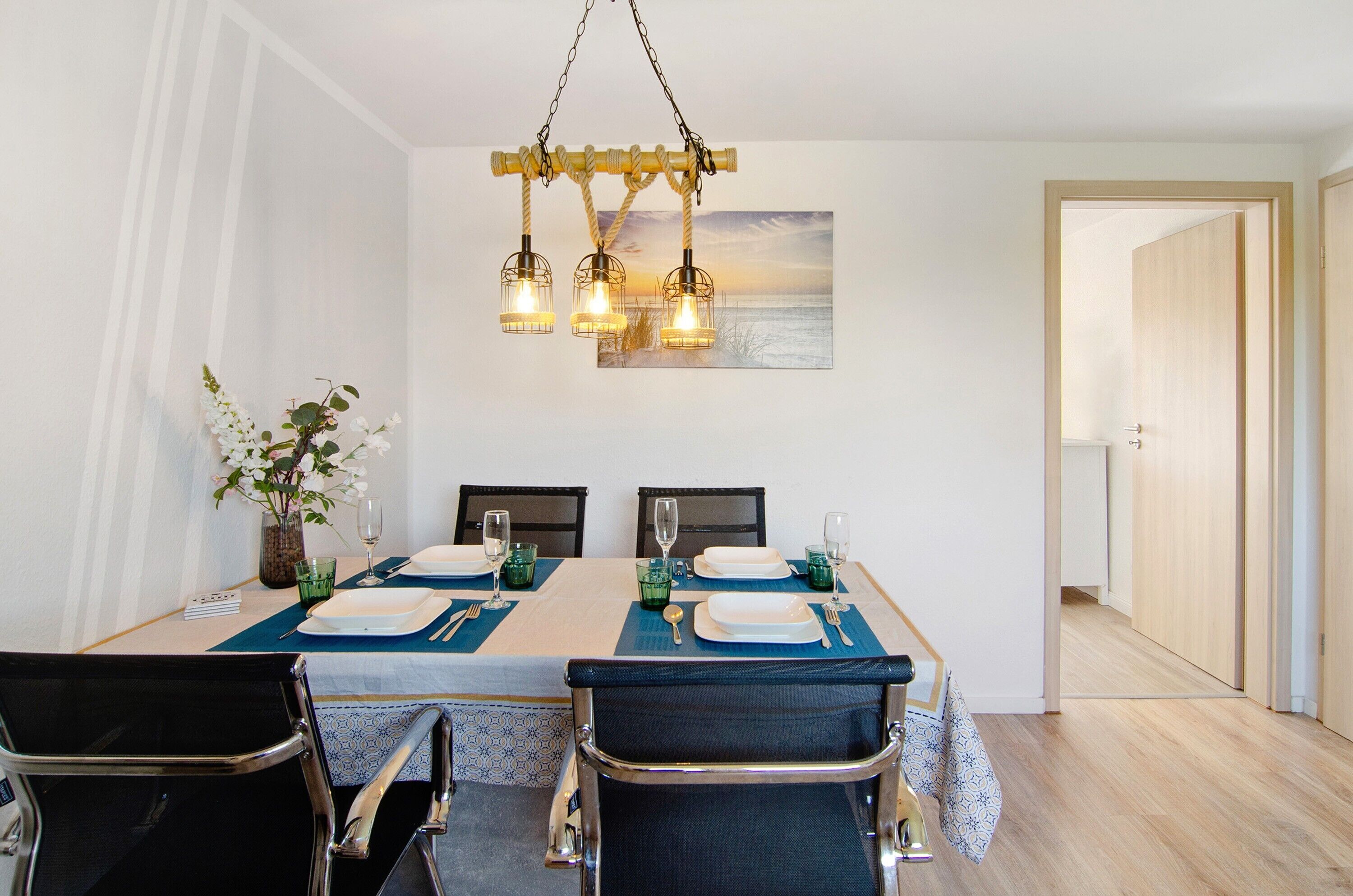 A dining area with a table set for four, chairs, a hanging light fixture, and a painting of a sunset over the ocean.