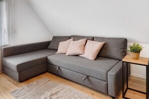 Un canapé d’angle gris avec trois coussins, une petite table d’appoint en bois sur laquelle est posée une plante, et un tapis beige sur un plancher en bois.