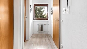 Un couloir doté d’un plancher en bois, d’un radiateur, d’une fenêtre donnant sur des arbres et d’une porte avec un interrupteur.