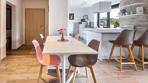 A modern kitchen with a wooden floor, a dining area with a white table and colorful chairs, and a view of the outdoors through a window.