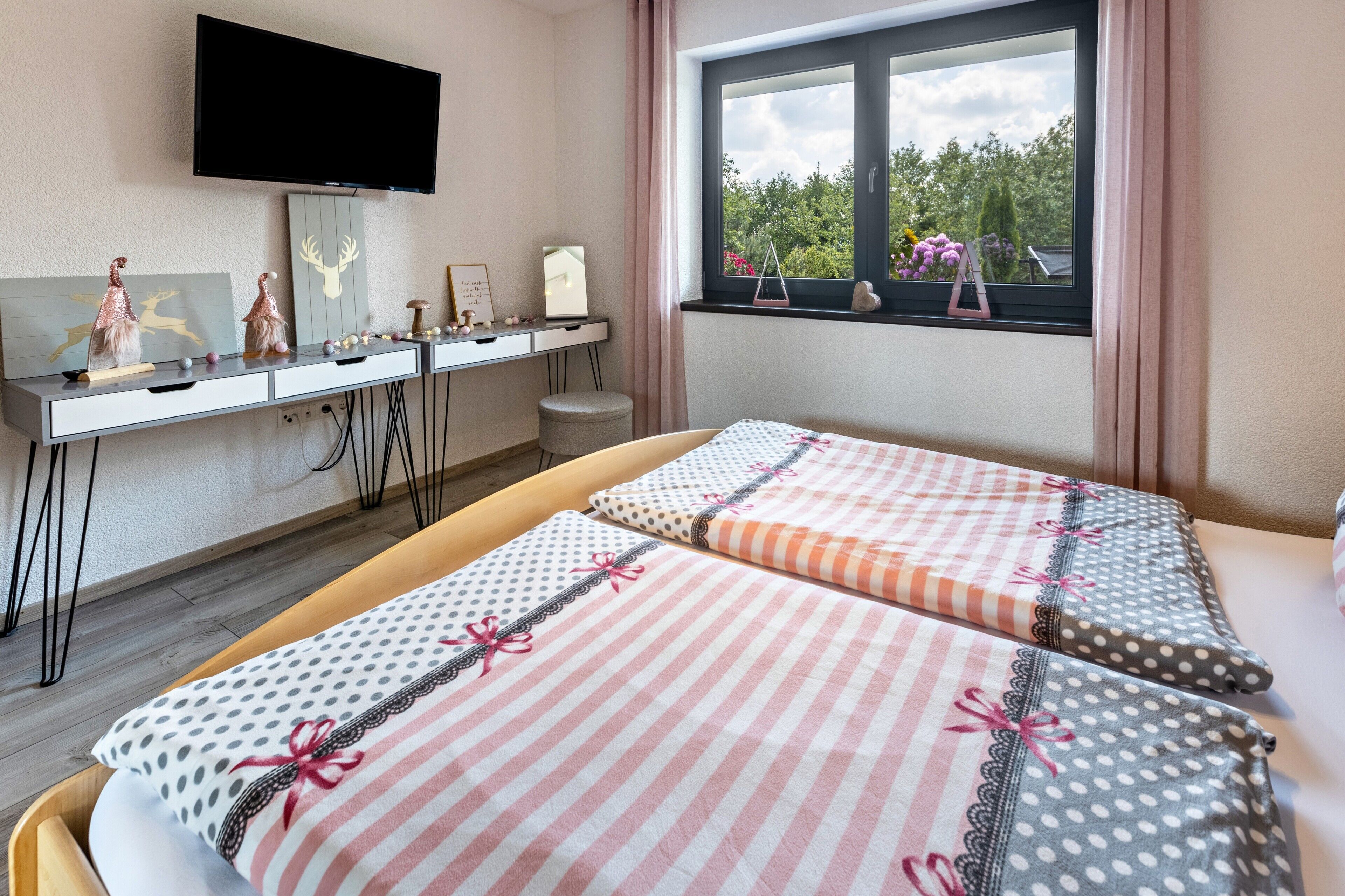 A hotel room with a bed, a desk with a flat-screen TV, a window with a view of greenery, and decorative items on the desk.