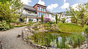 A modern house with a pond, surrounded by greenery and a clear sky.