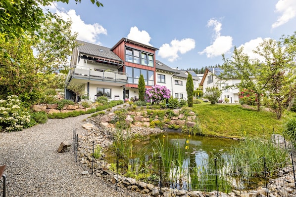 A modern house with a pond, surrounded by greenery and a clear sky.