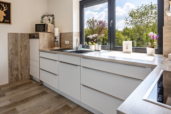 A modern kitchen with white cabinets, a black countertop, a sink, a microwave, and a window with a view of trees and sky.