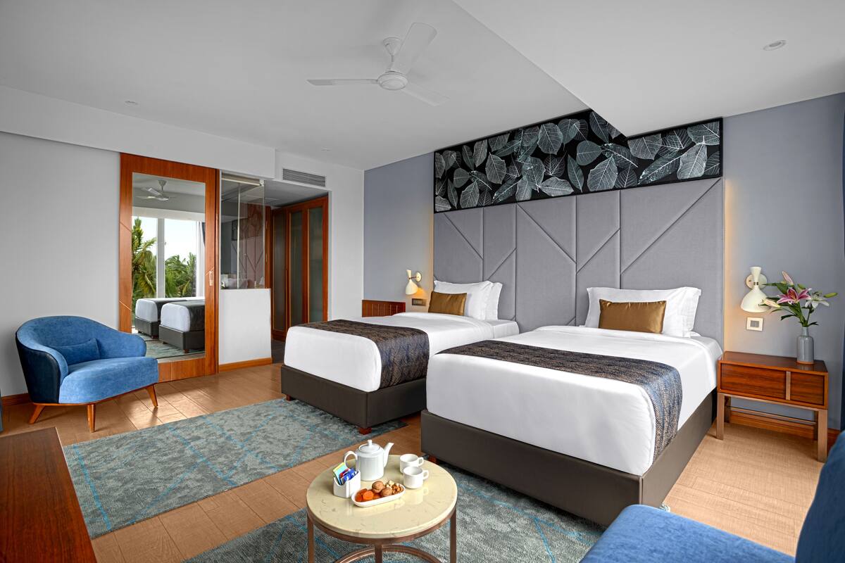 Una habitación de hotel moderna con dos camas, un sillón azul, una mesita redonda con provisiones para el desayuno y un panel decorativo en la pared.