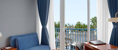 Une chambre d’hôtel avec un canapé bleu, une petite table ronde sur laquelle sont disposés des fruits, un bureau avec une chaise et un balcon offrant une vue sur les palmiers.