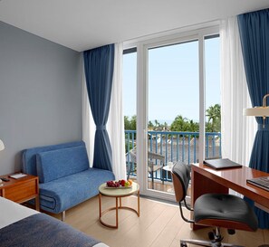 Habitación de hotel con sofá azul, una mesita redonda con fruta, escritorio con silla y balcón con vistas a palmeras.