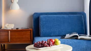 Una habitación con un sofá azul, una pequeña mesita de madera con un teléfono, una lámpara y una bandeja con uvas, una manzana y una naranja.