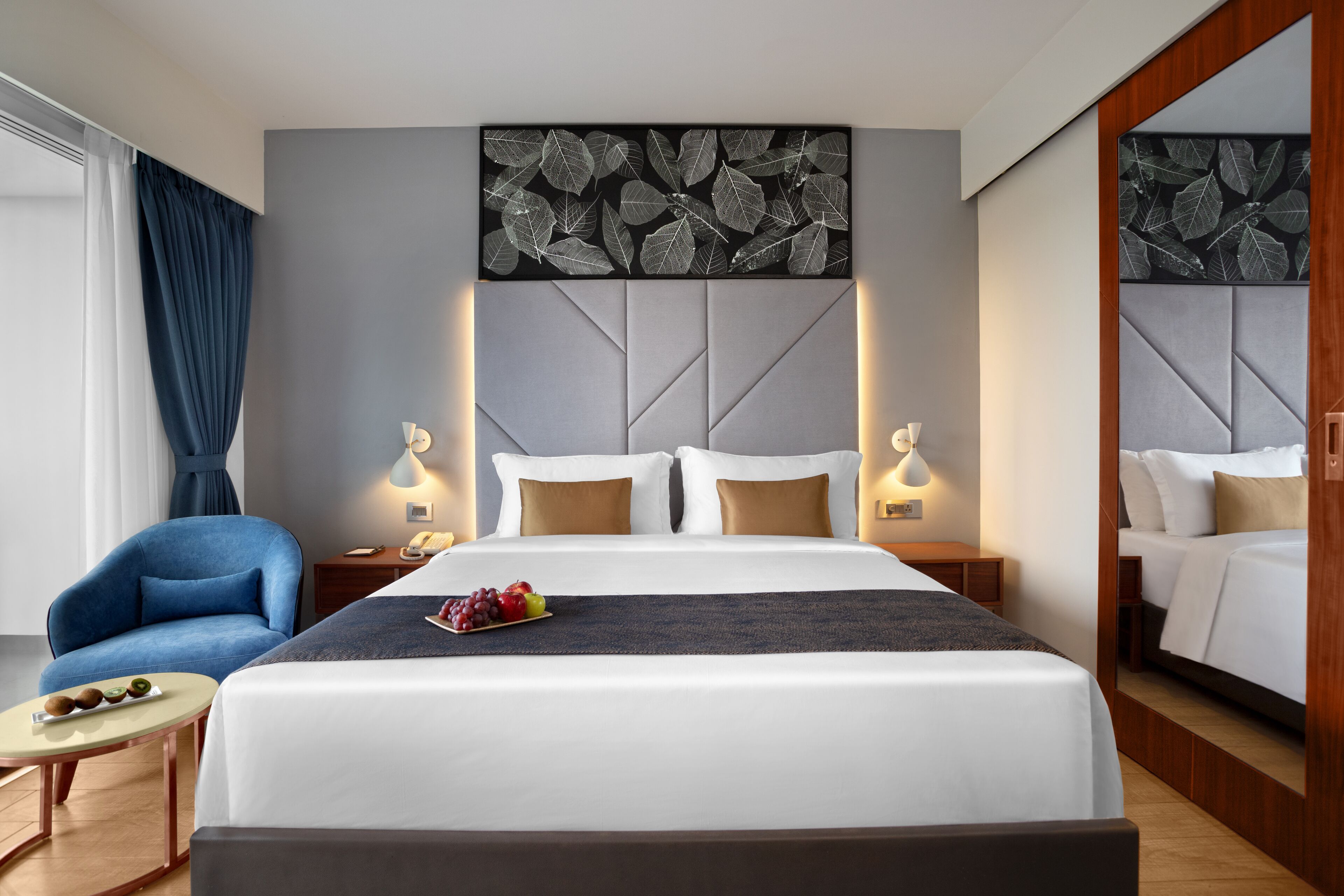 Una habitación de hotel moderna con una cama grande, un sillón azul, una mesita con fruta y una pared decorada con motivos de hojas.