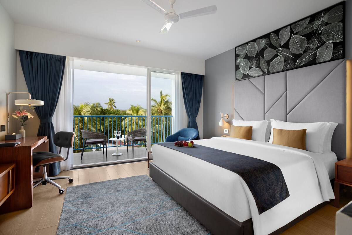 Una habitación de hotel moderna con una cama grande, un escritorio y un balcón con vista a palmeras.