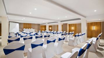 Une salle de conférence avec des rangées de chaises blanches ornées de rubans bleus, des murs lambrissés en bois et un tableau blanc sur le côté gauche.