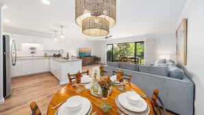 Een moderne keuken met witte kasten, een eetgedeelte met een houten tafel voor vier personen, en een grijze bank in de woonkamer.
