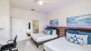 Habitación de hotel con dos camas, un escritorio y una silla. Se observa un ventilador de techo, un baño a través de una puerta abierta y obras de arte en la pared.