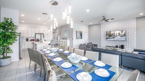 A modern kitchen with white cabinets, a dining area with a glass table set for six, and a living area with a gray sofa and a large TV on the wall.