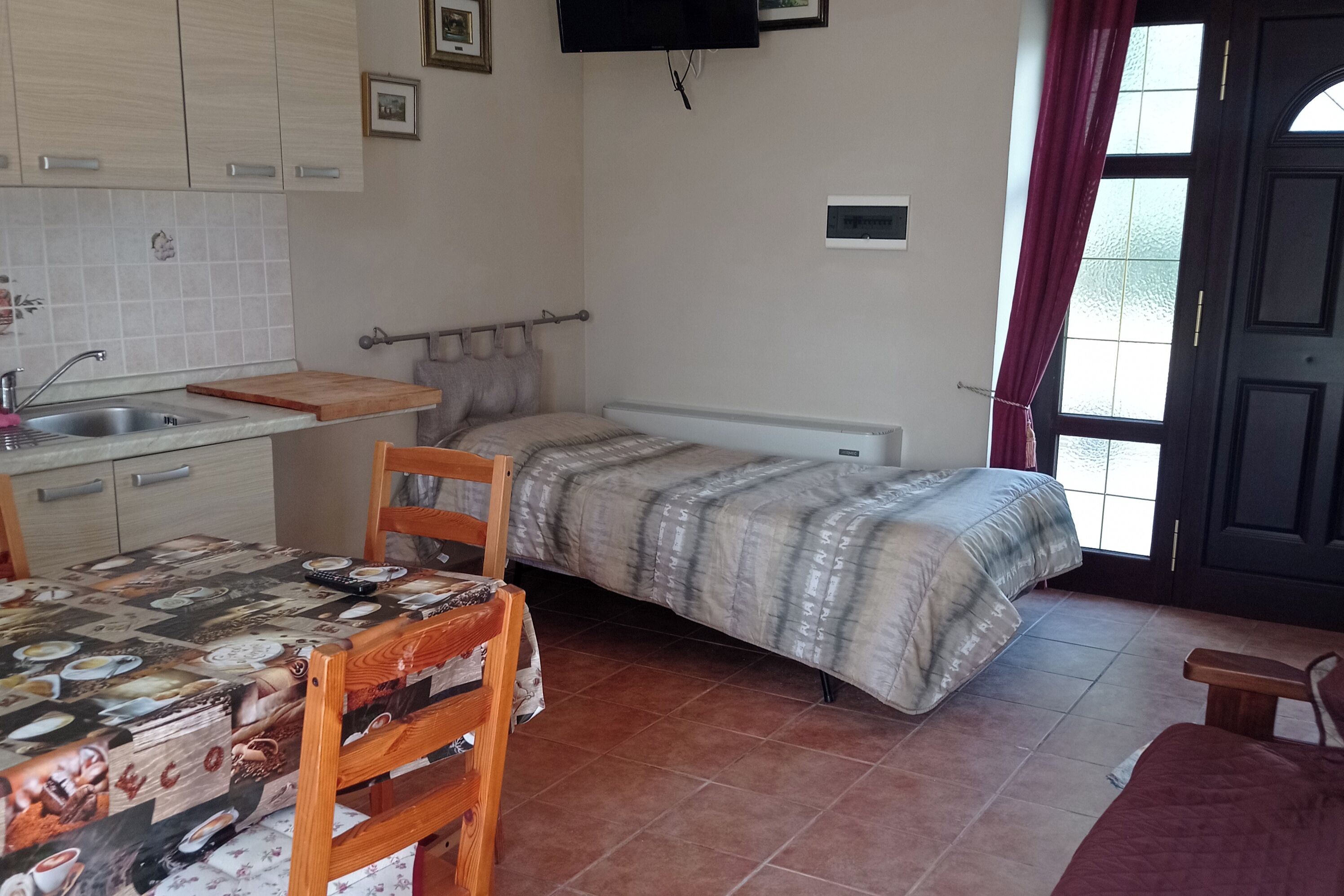 A compact room with a bed, a small kitchenette, a dining table, and a TV mounted on the wall.