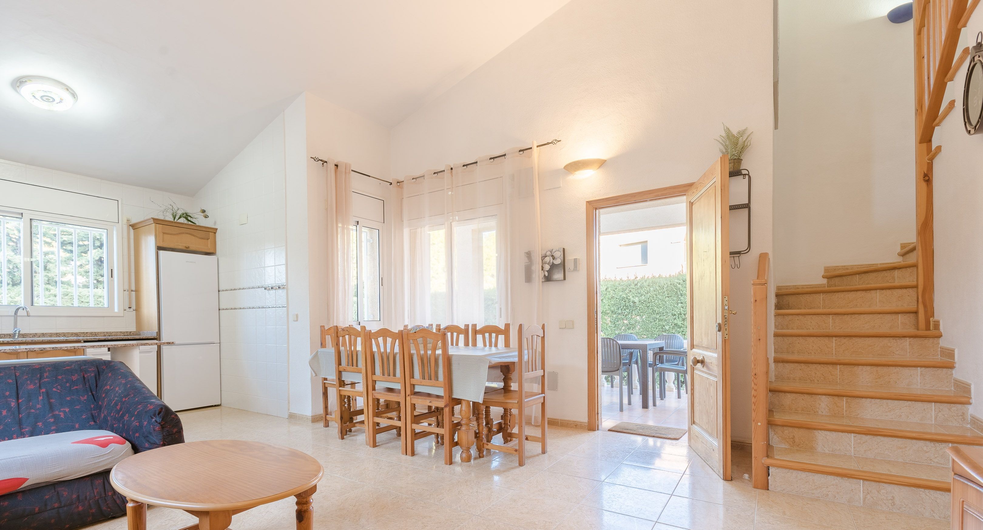 A spacious living area with a dining table and chairs, a sofa, a kitchen with a refrigerator, and a staircase.