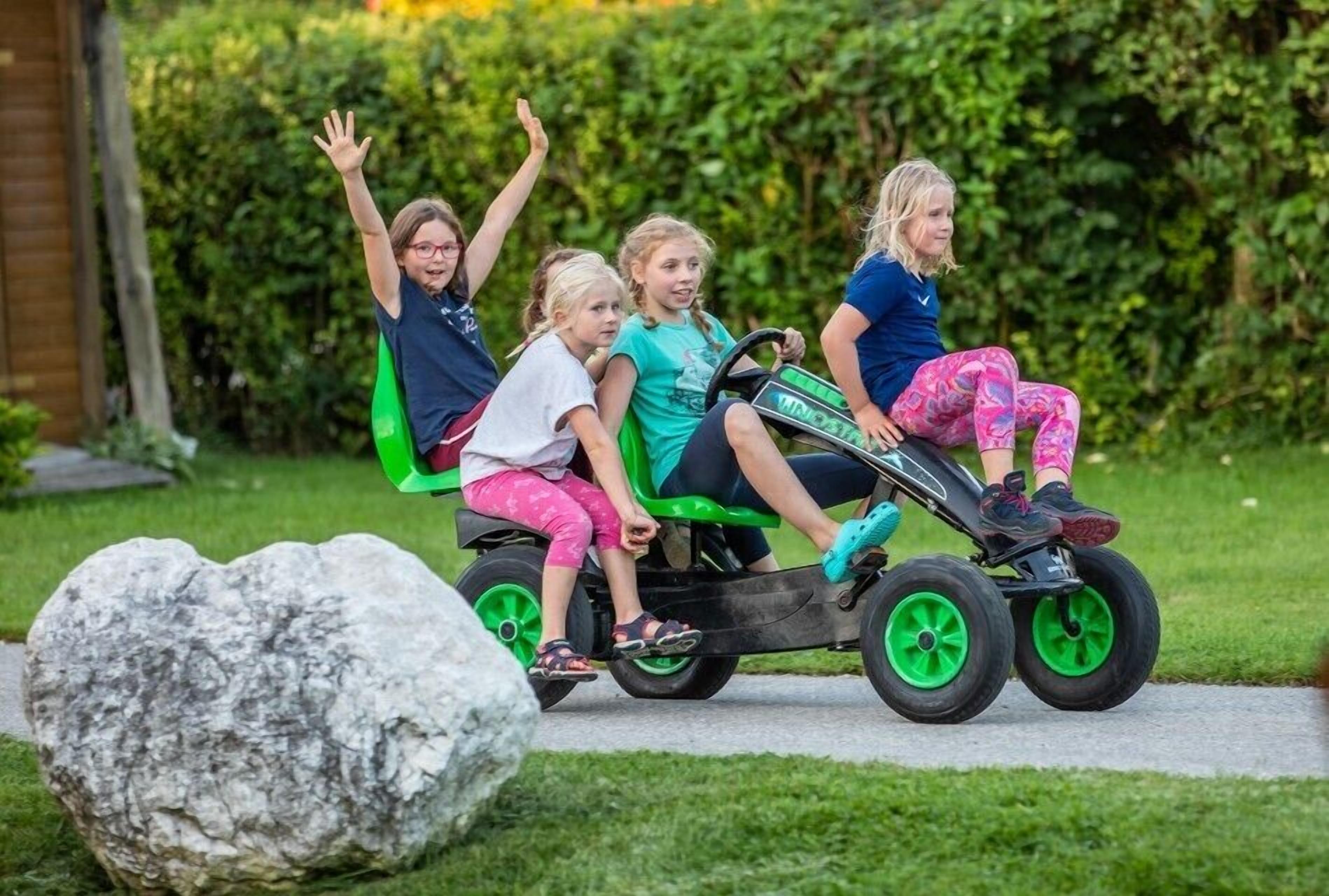 Vier Kinder auf einem grünen und schwarzen Bobbycar mit grünen Rädern, in einem Garten mit einem großen Felsen und einer Hecke im Hintergrund.