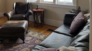 Un salon avec un canapé en cuir, un pouf assorti, une petite table en bois sur laquelle se trouve une lampe, et un tableau encadré accroché au mur.