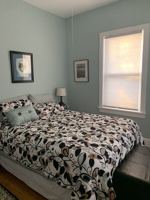 A bedroom with a bed, two framed pictures, a window with blinds, and a bedside table with a lamp.