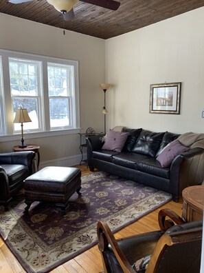 A living room with a dark leather sofa, a matching ottoman, a patterned rug, a wooden floor, a ceiling fan, and a window with a view of trees.