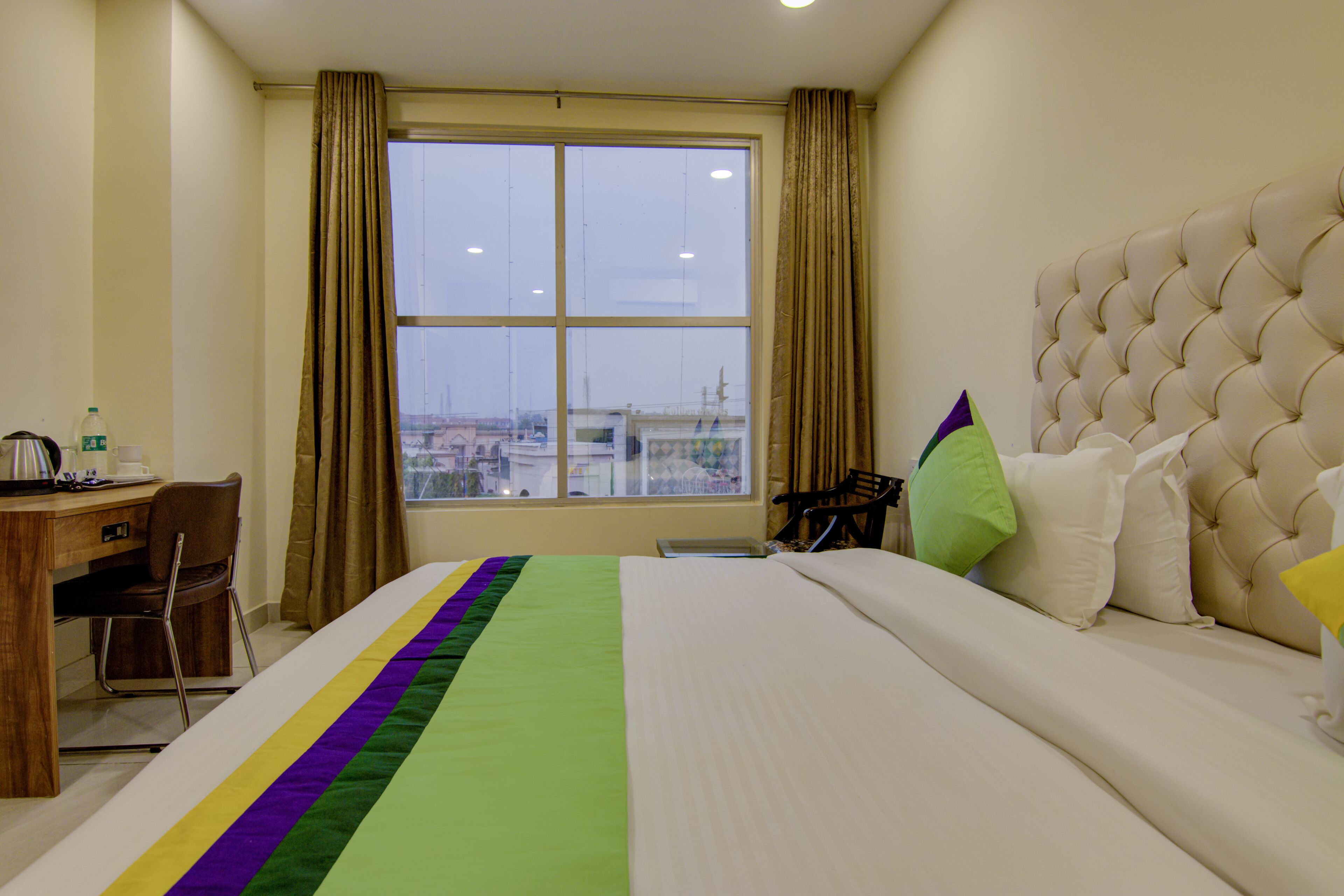 A hotel room with a large bed, a desk, and a chair. There is a window with curtains, and a view of the city.
