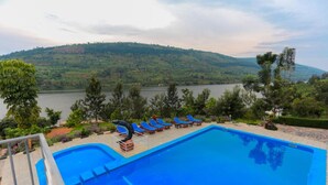 Une piscine à l’eau bleue, entourée de chaises longues, avec vue sur un lac et des collines en arrière-plan.