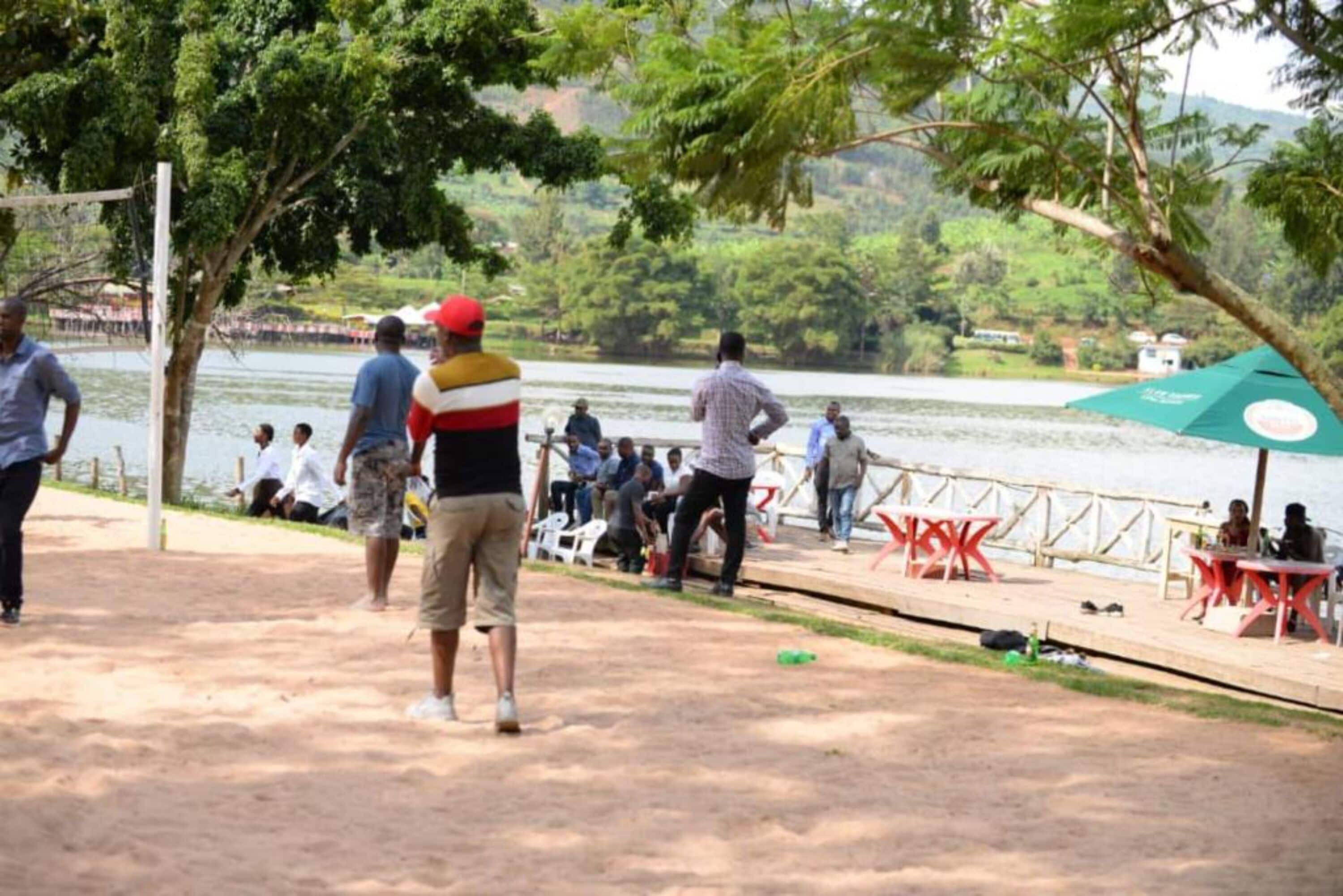 A lakeside park with people sitting and standing near the water, a volleyball net, and a gazebo.