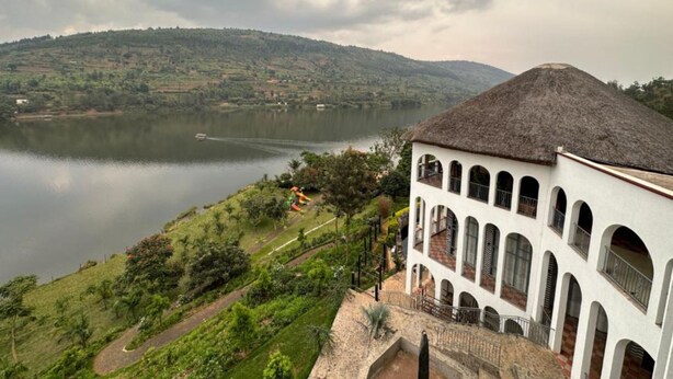 Un bâtiment blanc, doté de fenêtres en arc de cercle et d’un toit de chaume, surplombe un lac paisible et un paysage vallonné.