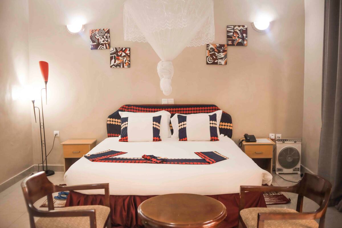A hotel room with a large bed, two bedside tables, a standing heater, a chair, and a small round table.