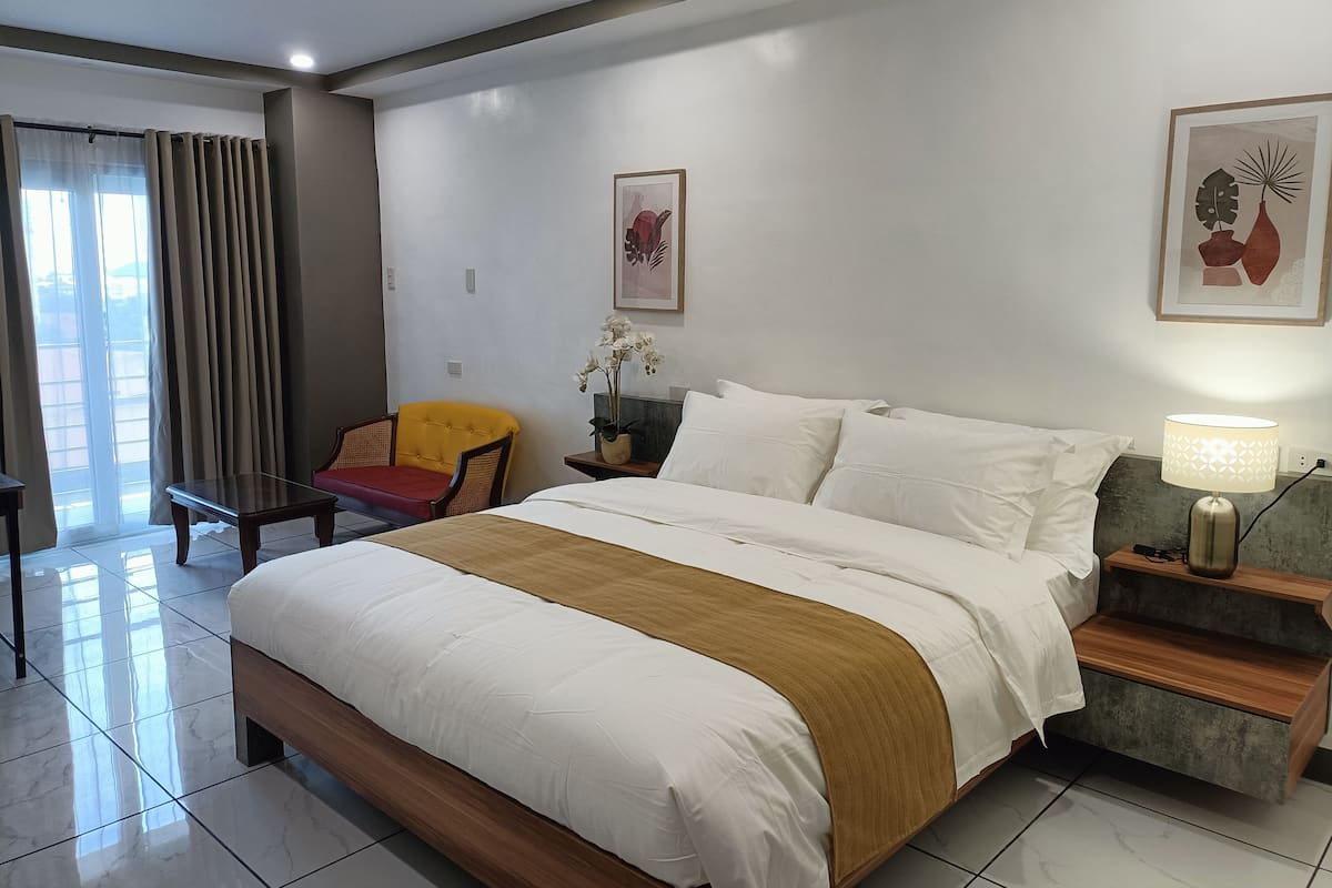 A hotel room with a large bed, a nightstand with a lamp, a chair, and a window with curtains.