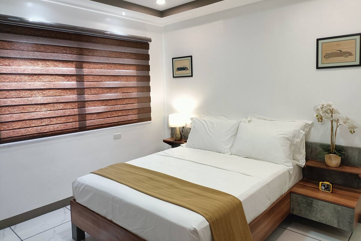 A neatly made bed with white linens and a brown throw, a bedside table with a lamp, a framed picture on the wall, and a window with blinds.