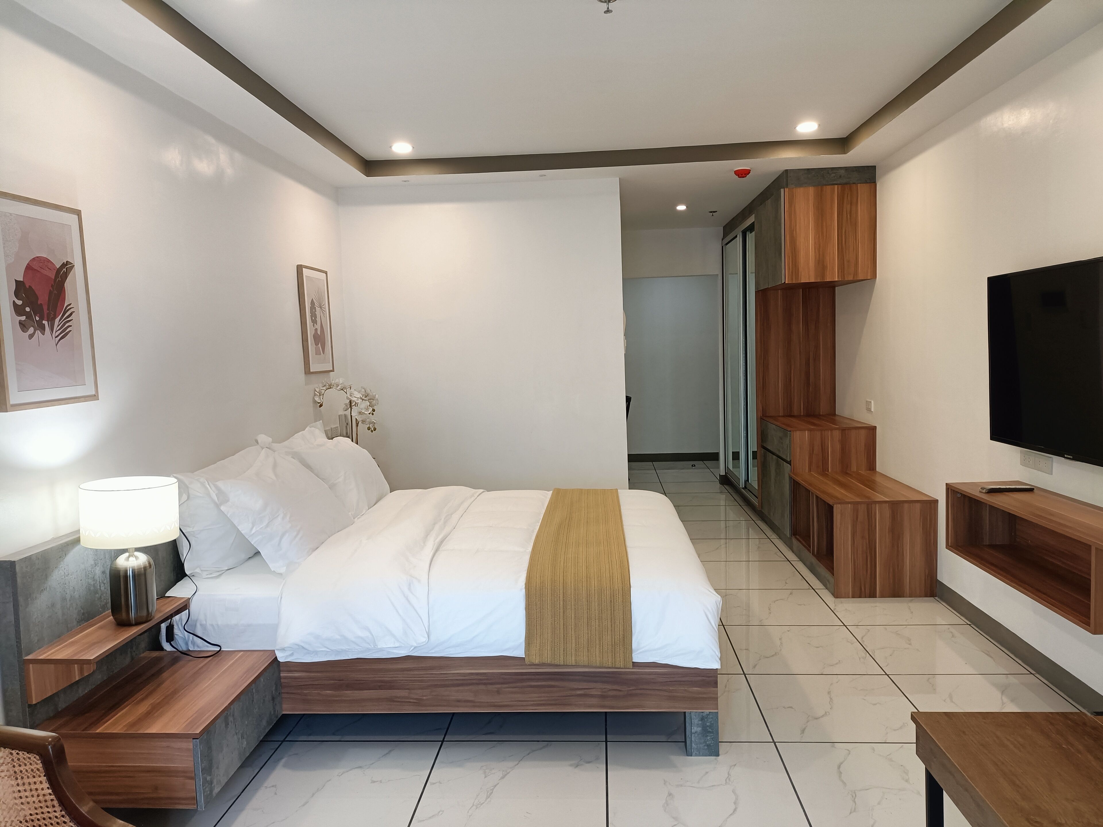 Una habitación de hotel moderna con una cama grande, mesitas de noche de madera, un televisor y un pasillo que conduce a un baño.