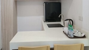 Une cuisine de taille réduite avec un plan de travail blanc, deux chaises en bois, un réfrigérateur noir et une bouilloire.