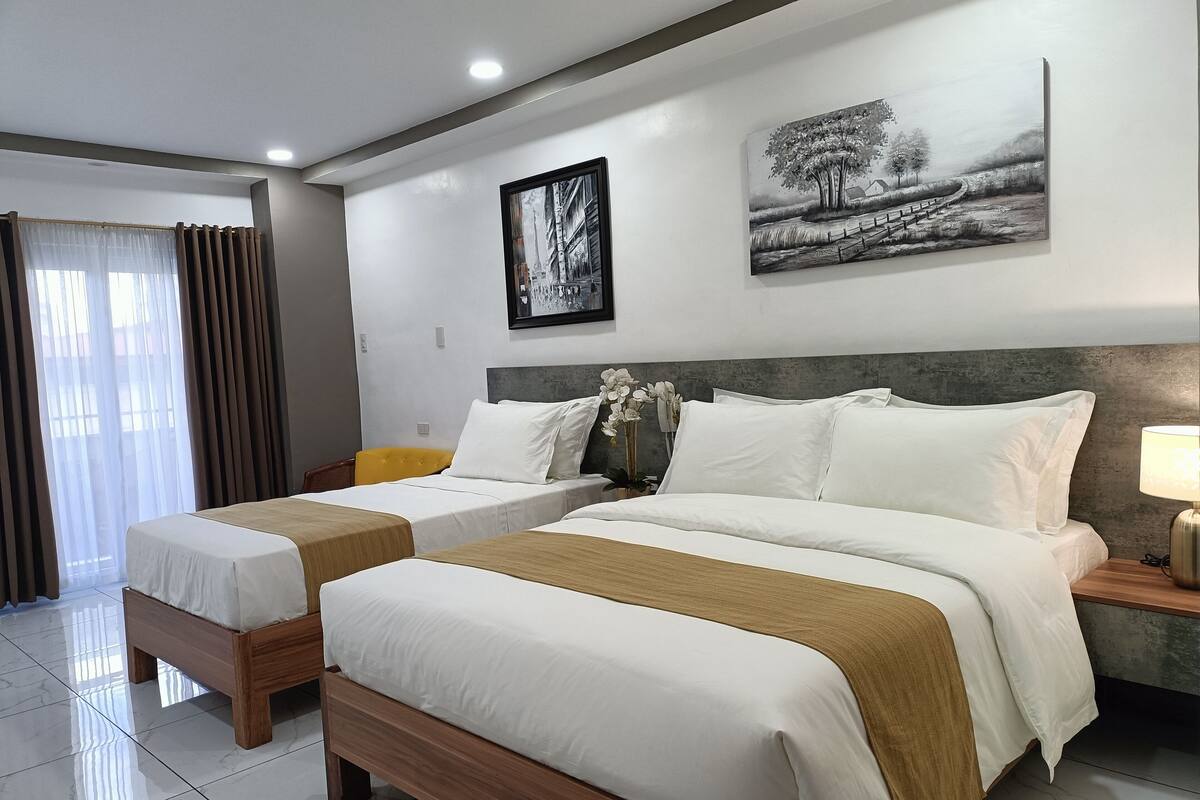 A hotel room with two beds, a large window with curtains, a painting on the wall, and a nightstand with a lamp.