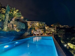 Une vue nocturne d’un hôtel avec une piscine, un escalier menant à l’entrée et de la verdure environnante.