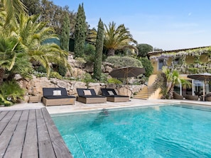 A poolside area with lounge chairs, a wooden deck, and a stone wall with vegetation.