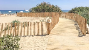 Une promenade en bois mène à une plage de sable, où l’on trouve des parasols et d’où l’on a une vue dégagée sur la mer.