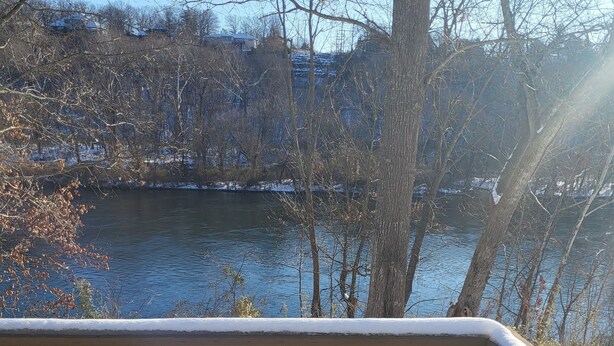 A wooden deck with a snow-covered bench, overlooking a river with trees and buildings in the background. - Lakeside, fishing, Cozy 2/2 - 5 minutes from Branson attractions and Table Rock (Branson)