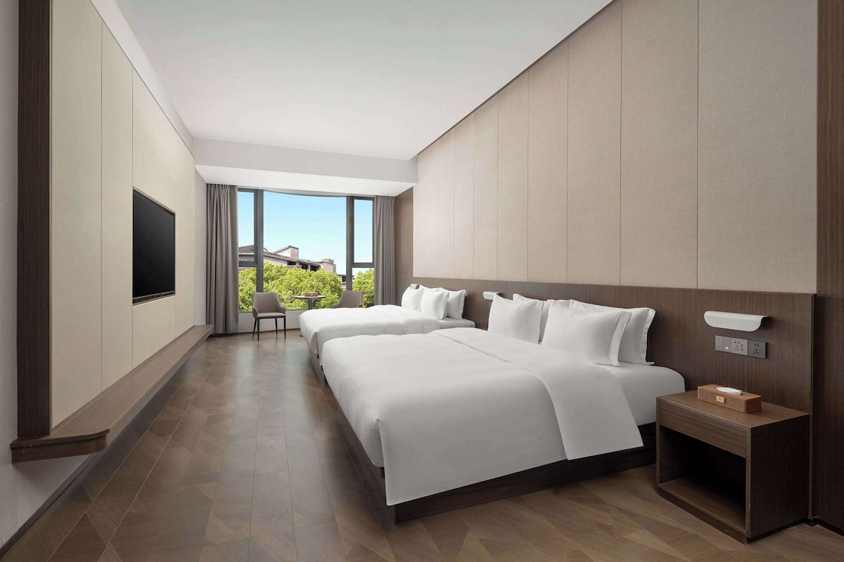 Quarto de hotel moderno com duas camas, uma televisão, uma mesa de cabeceira e vista para o verde lá fora.
